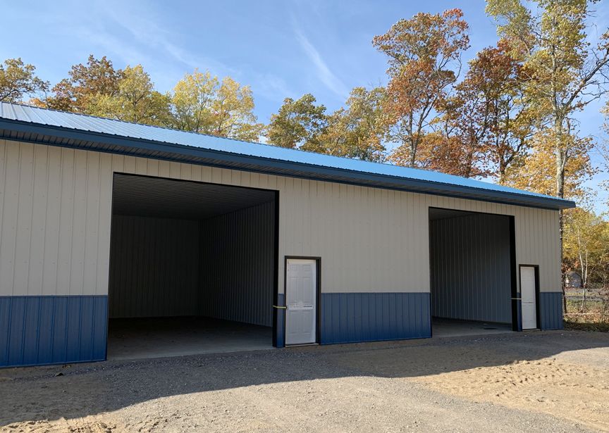 White metal garage building with blue trim, open bay doors, and a gravel lot under autumn trees