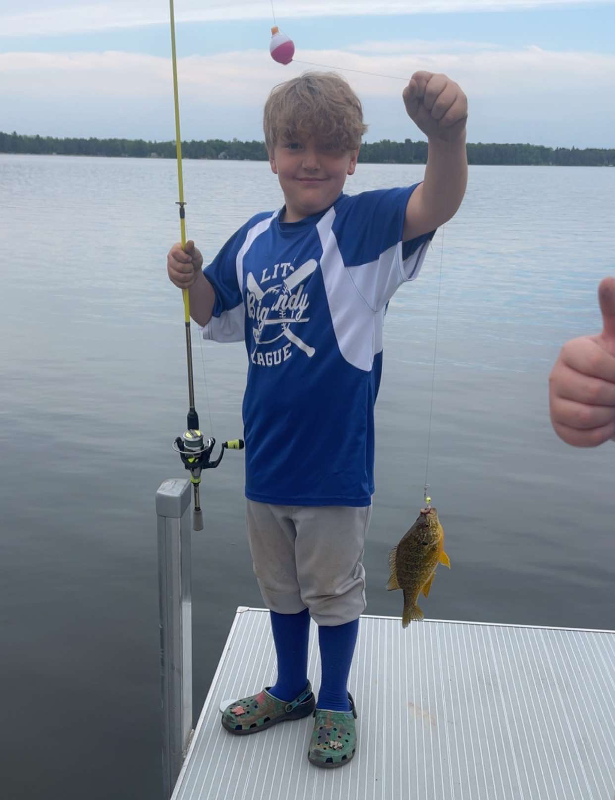 Child on a dock holding a fishing rod, proudly showing a small fish by a calm lake.