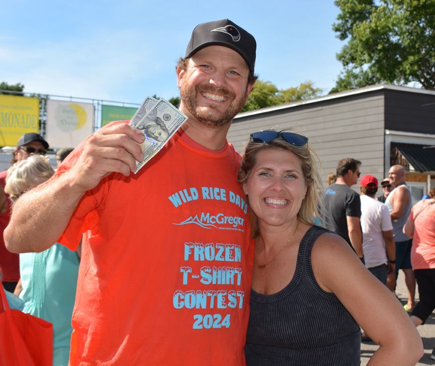 Smiling pair at outdoor event; man in orange shirt holds cash and woman stands beside him.