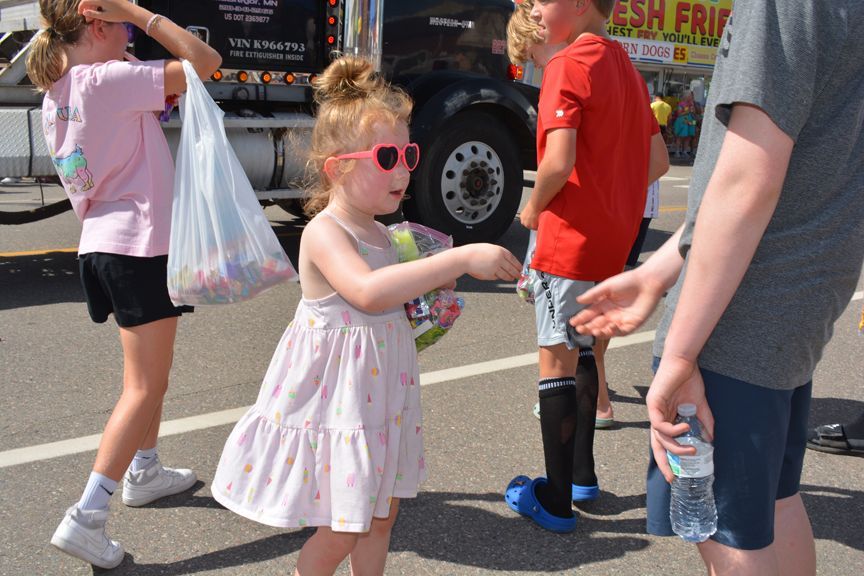 Children at an outdoor festival, one in pink sunglasses holding a hand with a water bottle nearby.