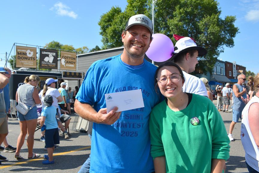 Two smiling people posing outdoors at a sunny event, one holding a white paper sign.