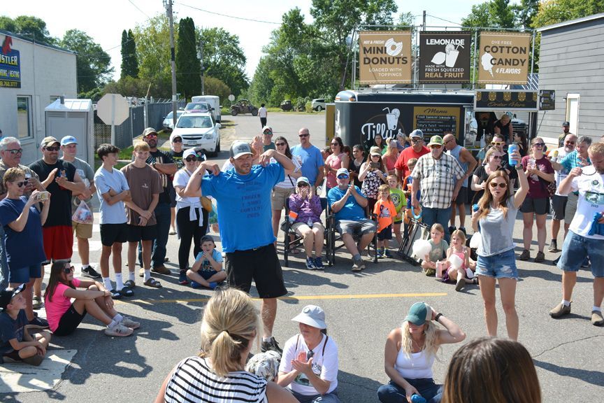 Crowd gathered outdoors at a fair, watching a man in a blue shirt perform in a parking lot