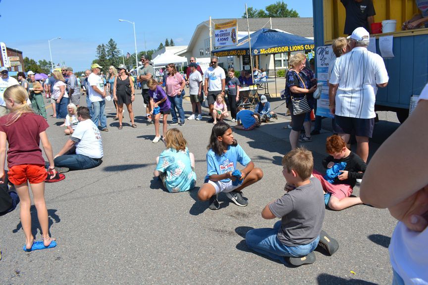 Children and adults gathered outdoors at a street fair, with kids sitting and playing on the pavement.