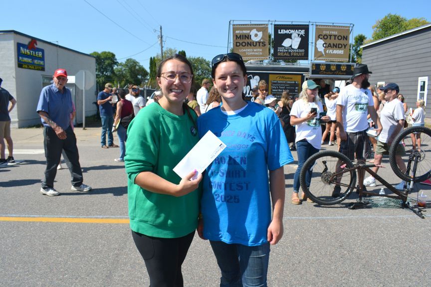 Two people posing outdoors in front of a crowd at a sunny street event, one holding a folder.