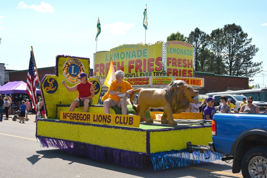 Lions Club parade float with lemonade and fries signs, lion figures, and a blue truck in a sunny street scene