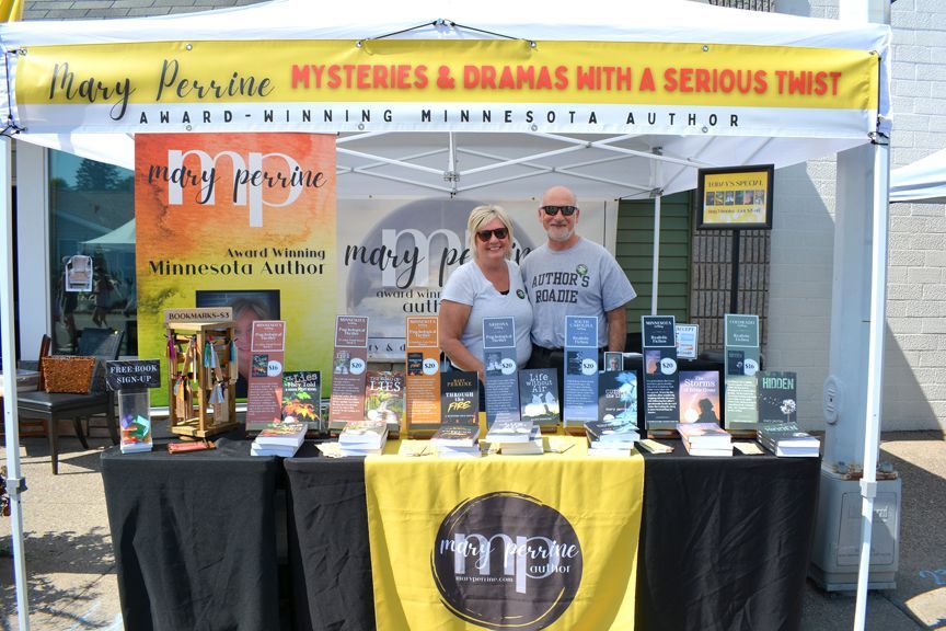 Authors at a book fair booth under a white tent, with colorful mystery novels and a yellow tablecloth.