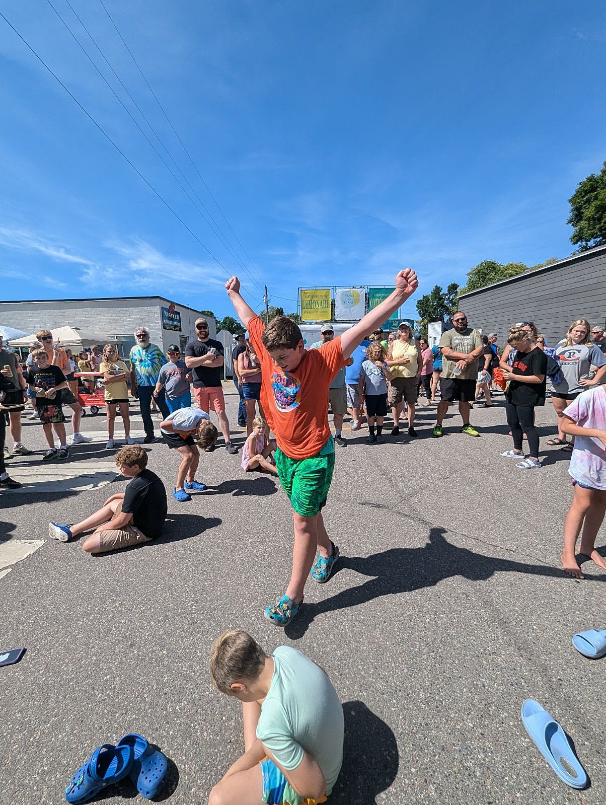 People gathered outdoors; a person in orange shirt and green shorts stands with arms raised at center.