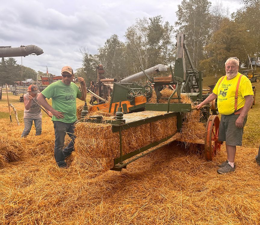 People standing by a farm machine in a woodchip-covered field, with logs and equipment in the background.