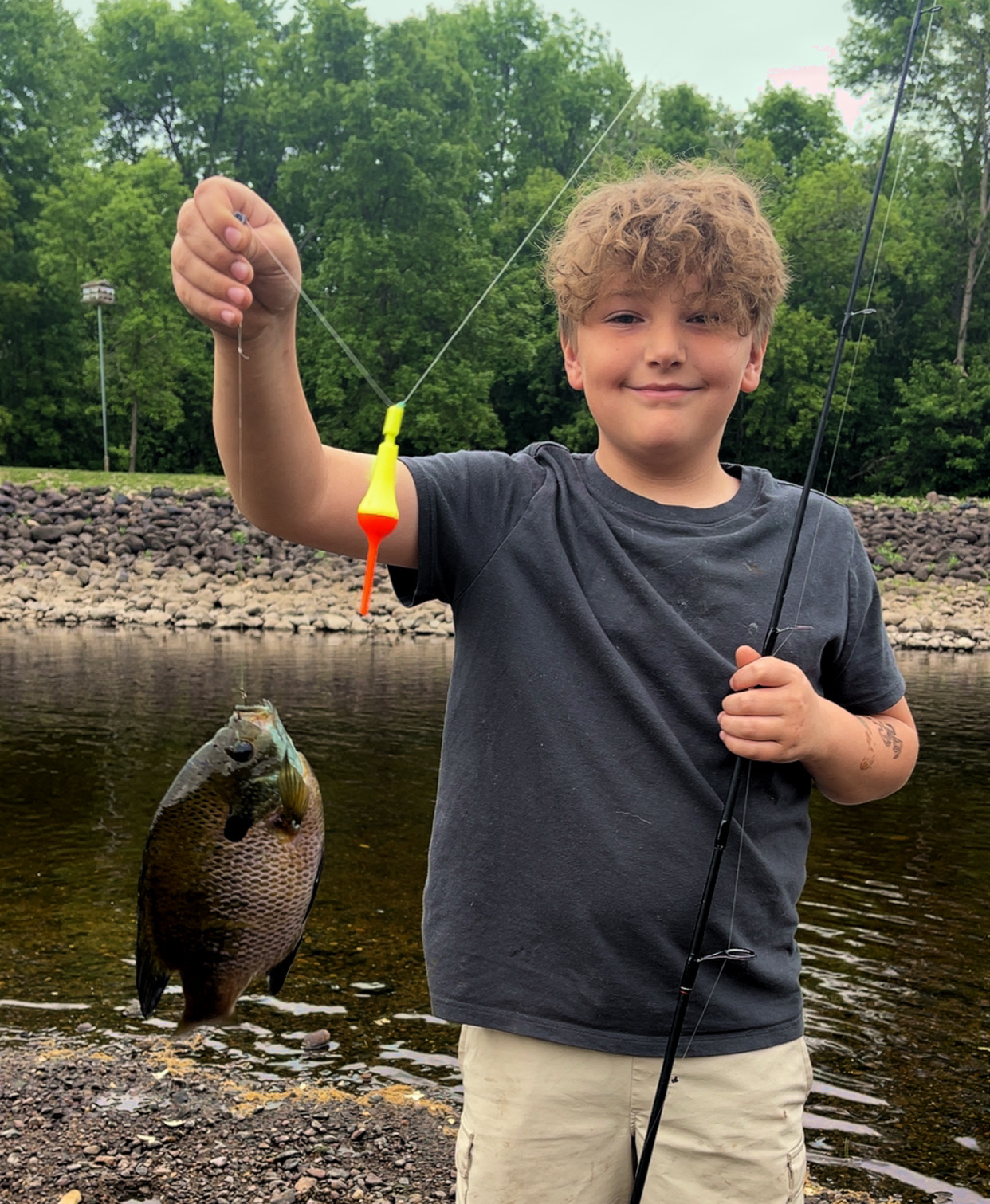 Child smiling by a river, holding a fishing rod with a colorful lure and a caught fish.