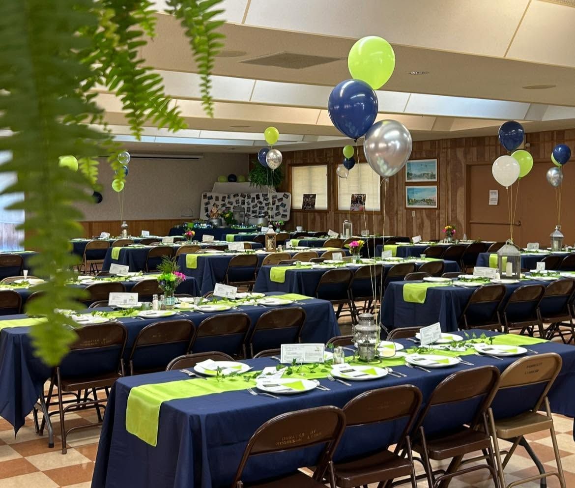 Banquet hall set with round tables, blue chairs, green napkins, and blue-silver balloons