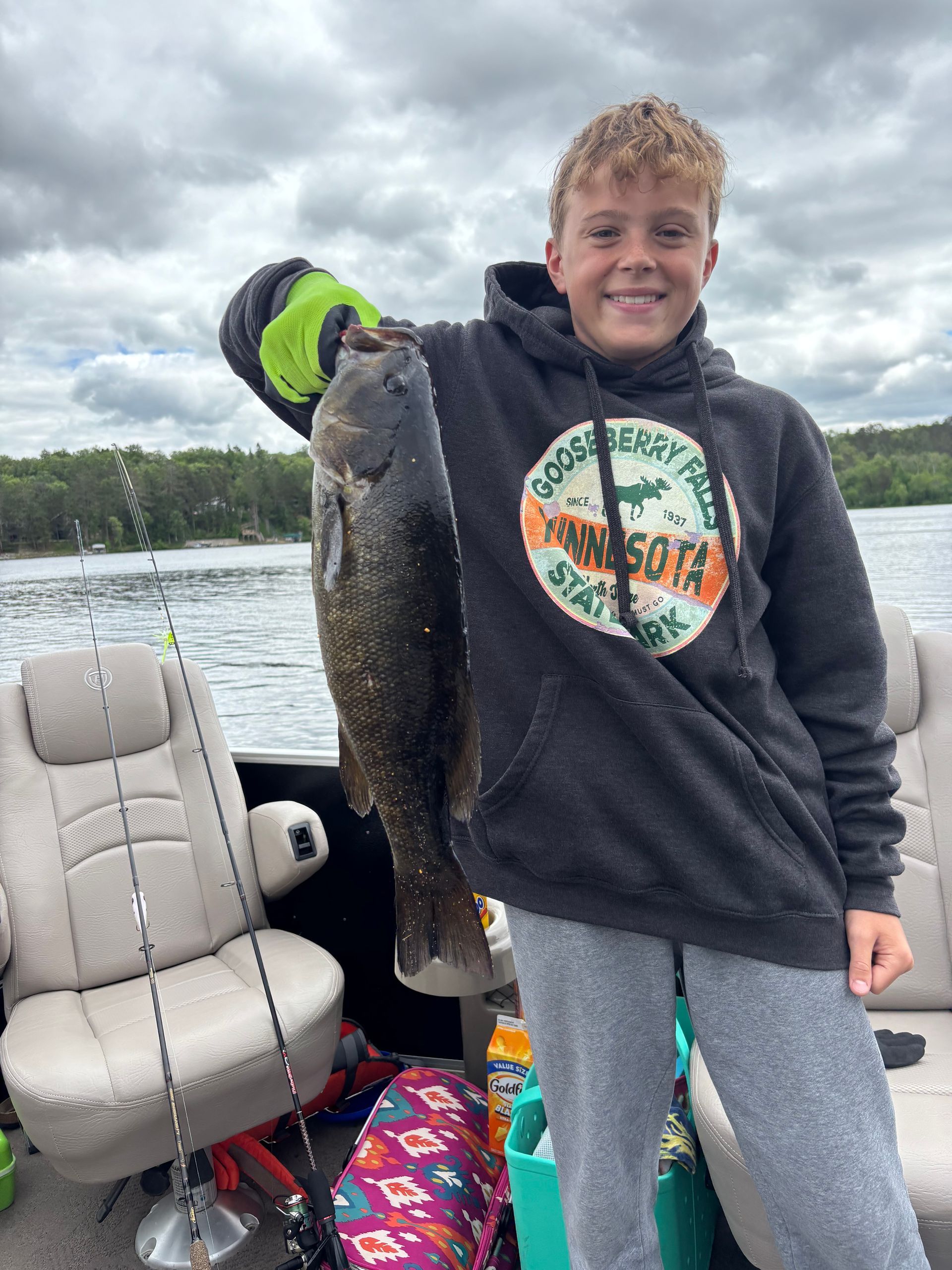 Person holding a large fish on a boat, wearing a dark hoodie and neon glove, with cloudy water and shoreline behind.