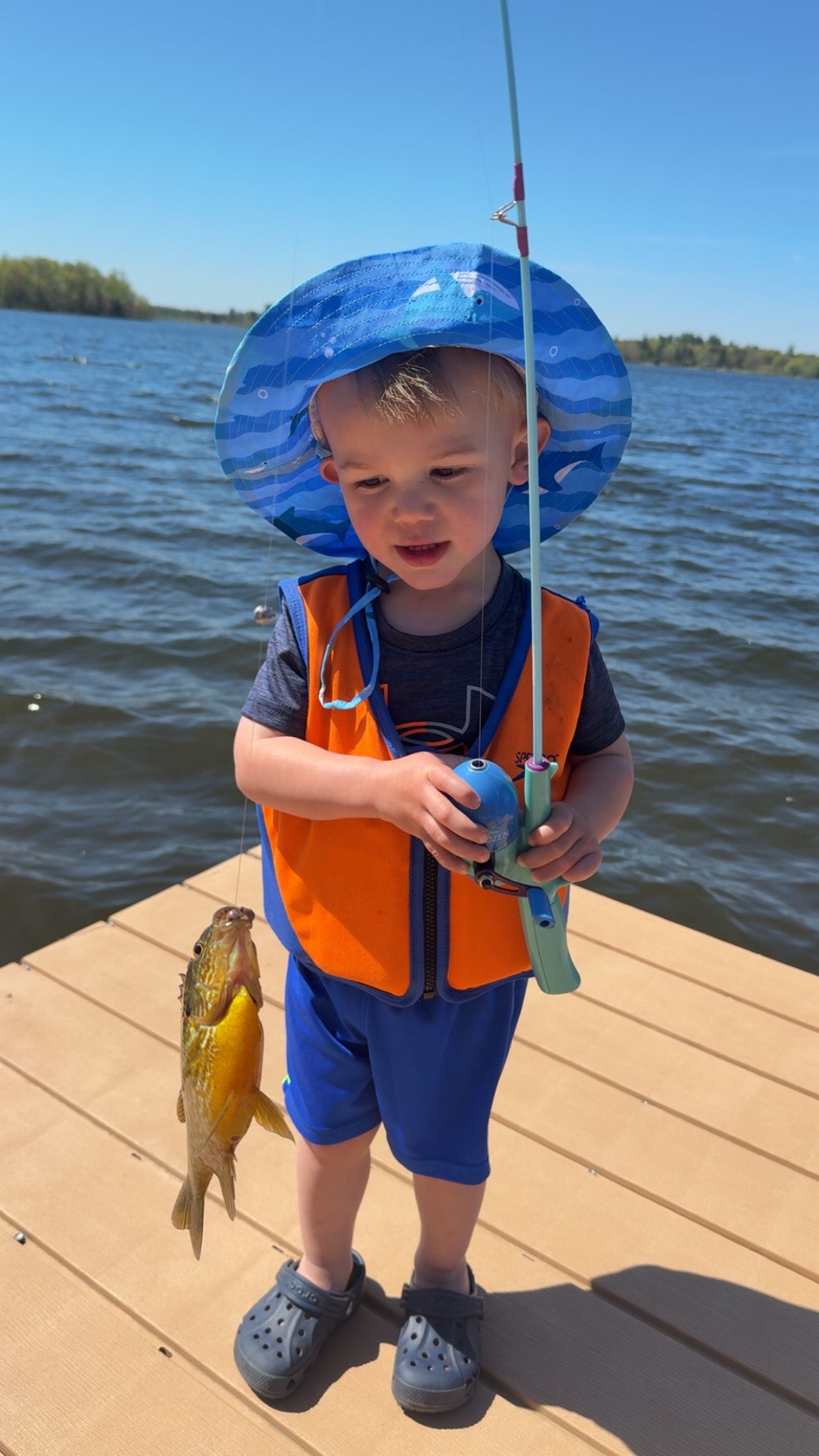 Child in orange life jacket and blue sun hat holding a fishing rod on a dock by the water.