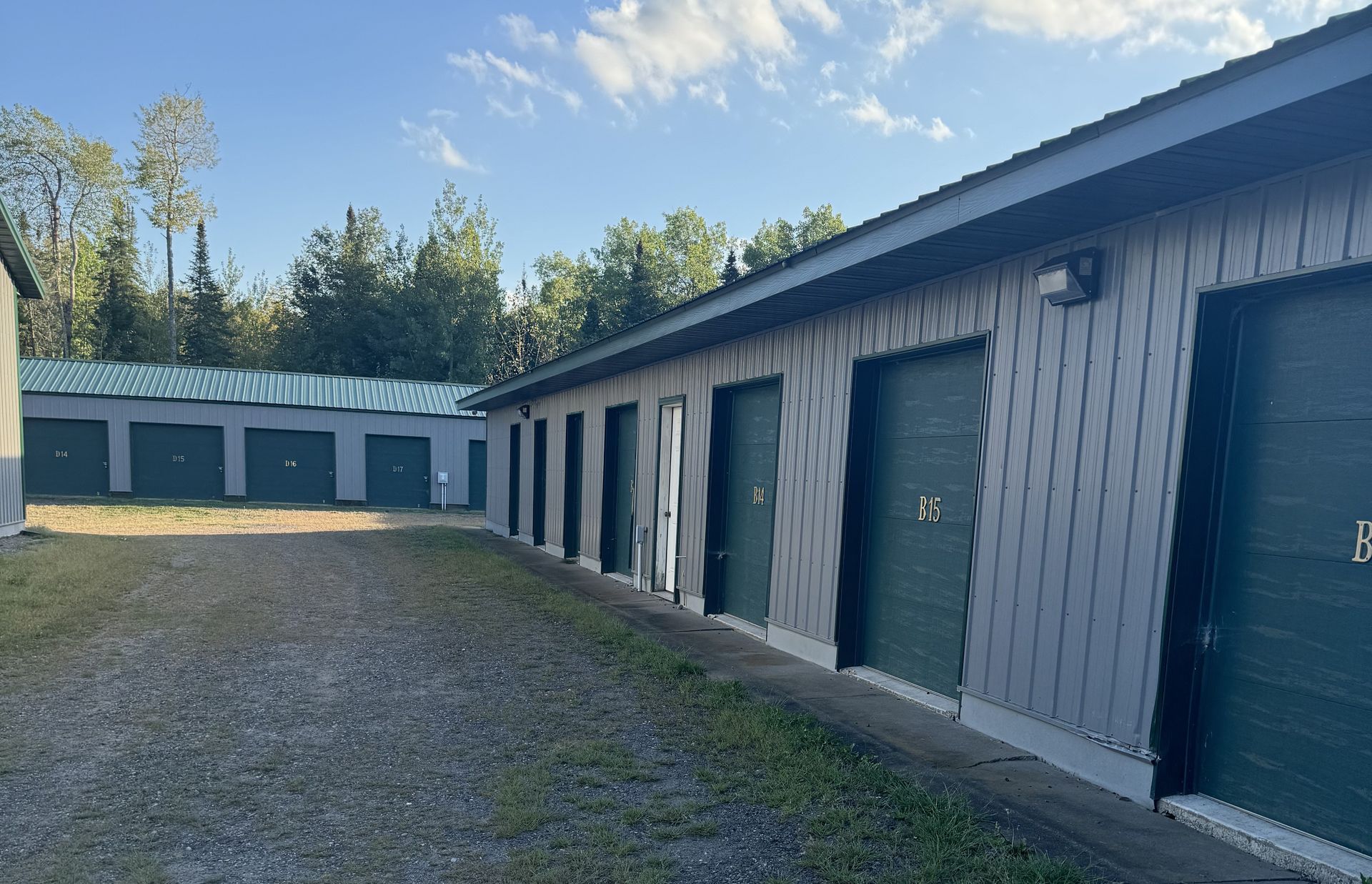 Row of gray storage unit garages with green doors beside a gravel driveway under a clear sky