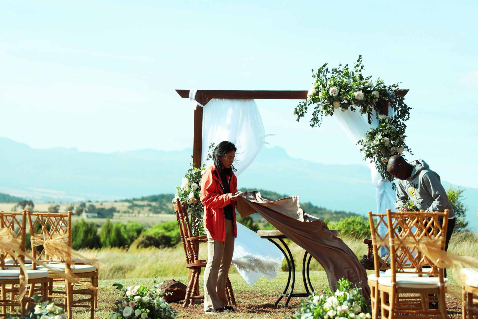 Bride in red stands by a floral wedding arch with chairs, overlooking a sunny mountain landscape.