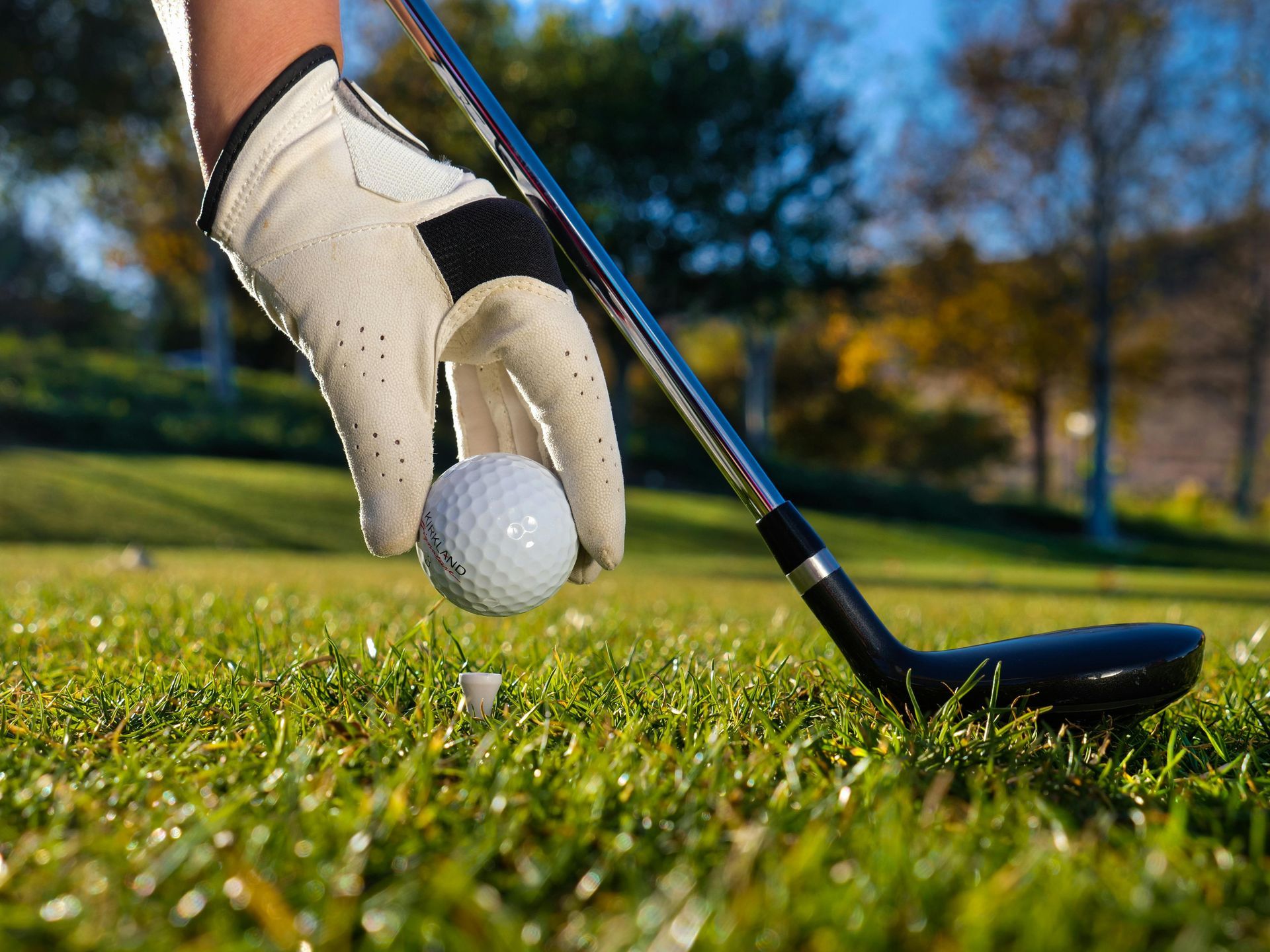 Golfer’s gloved hand tees up a golf ball beside a club on a grassy course.