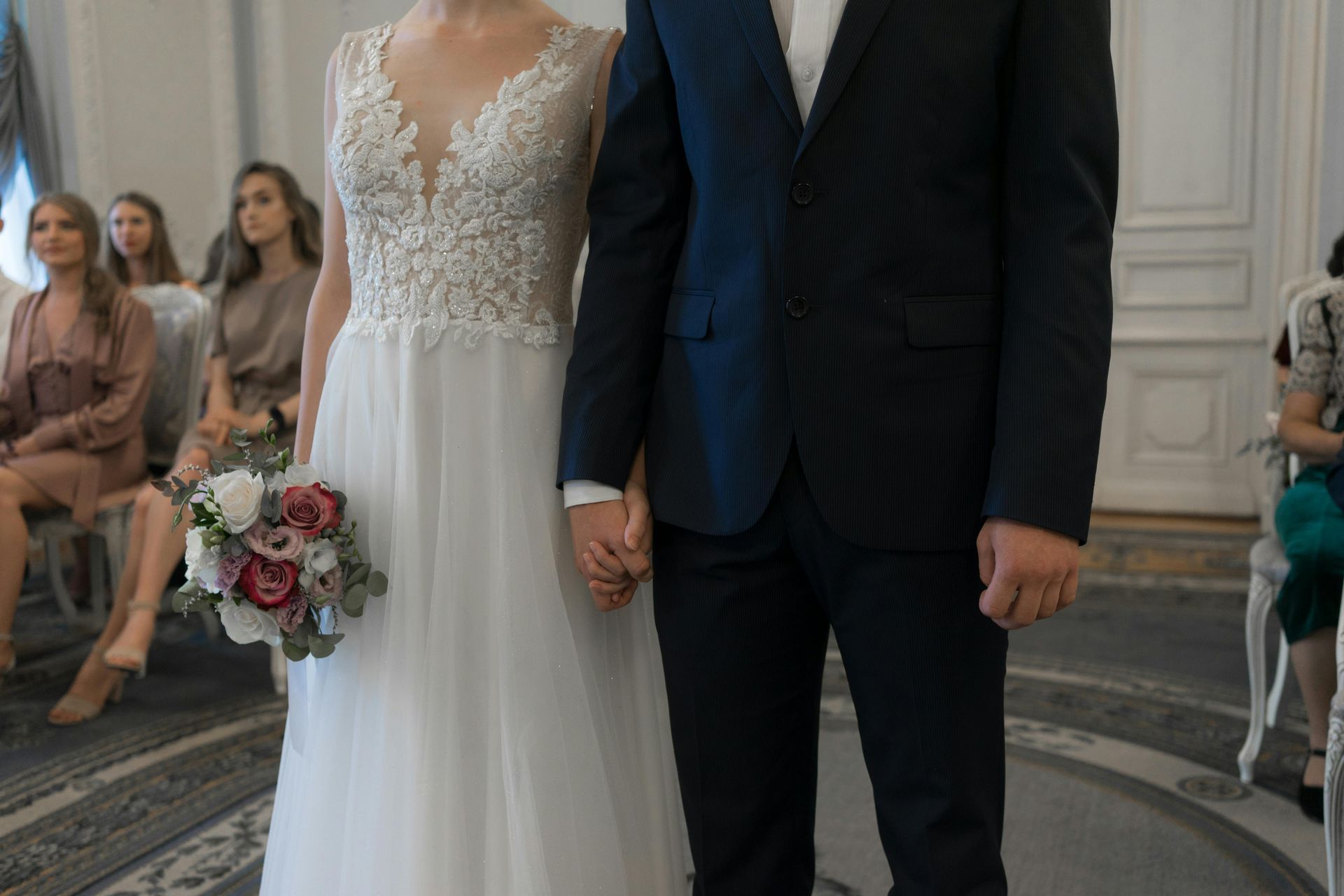 Bride and groom holding hands at a wedding ceremony, with guests seated in the background.
