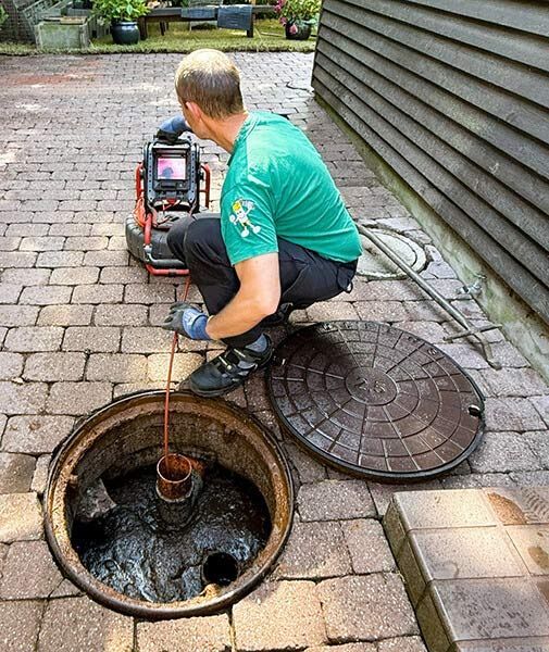 Worker inspecting sewer line with camera near open manhole on brick driveway
