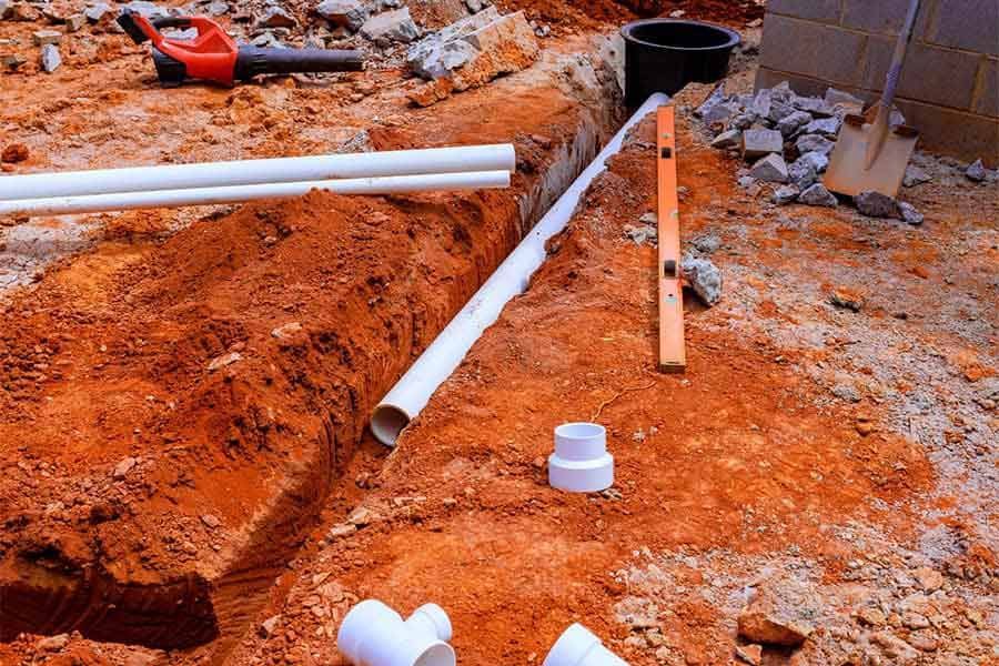 Construction trench with white PVC pipes laid in orange dirt and rubble, with tools nearby
