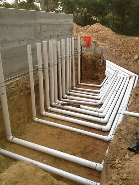 White PVC pipes run in a trench beside a concrete wall at a construction site.