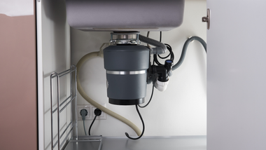 Under-sink view of a garbage disposal with plumbing, wiring, and hoses inside a cabinet