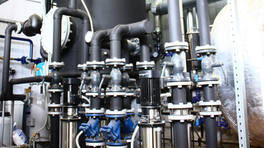 Industrial piping and valves in a mechanical room with black and blue pipes and metal tanks