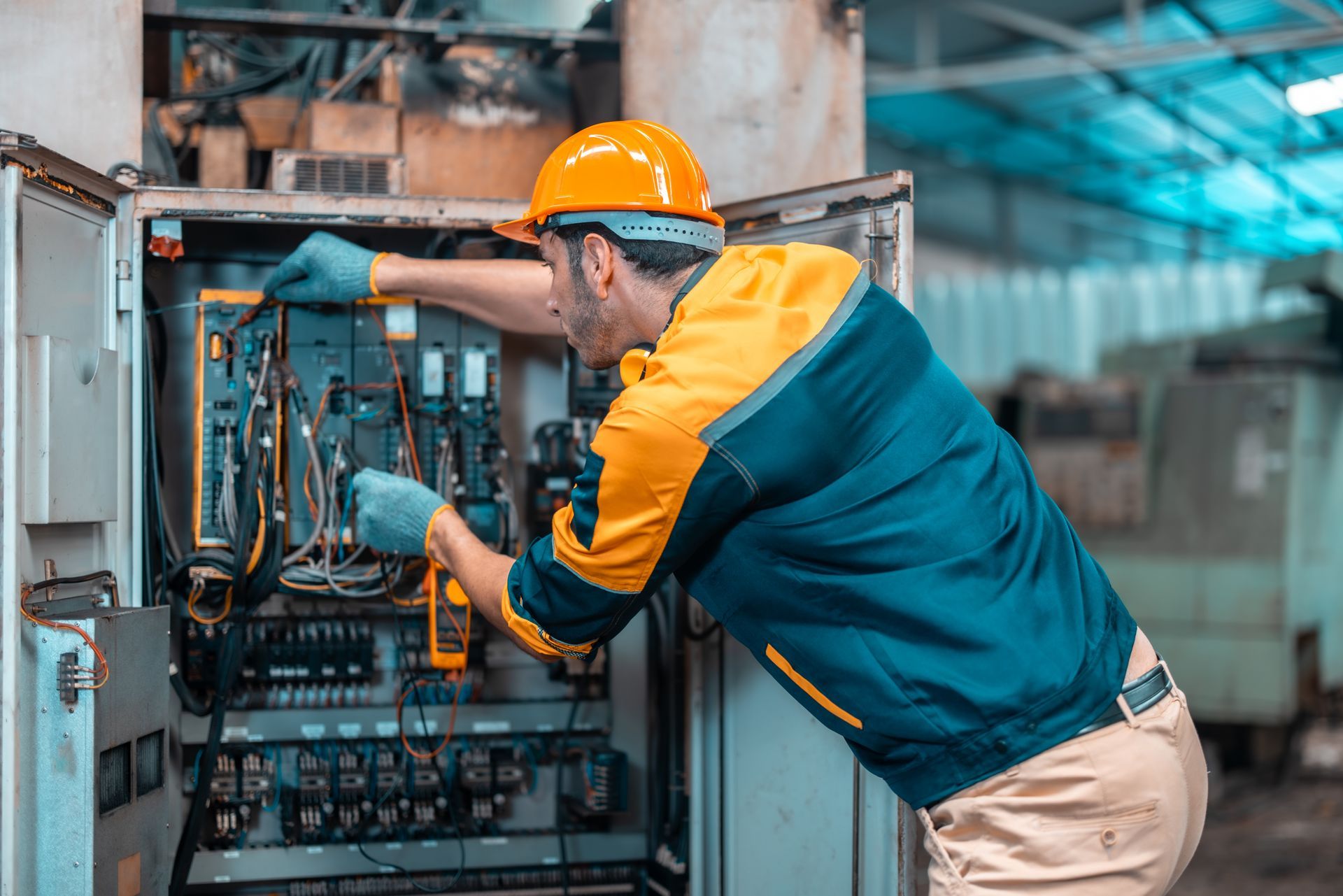 A technician in a hard hat and protective gloves inspects electrical wiring inside an industrial control panel.