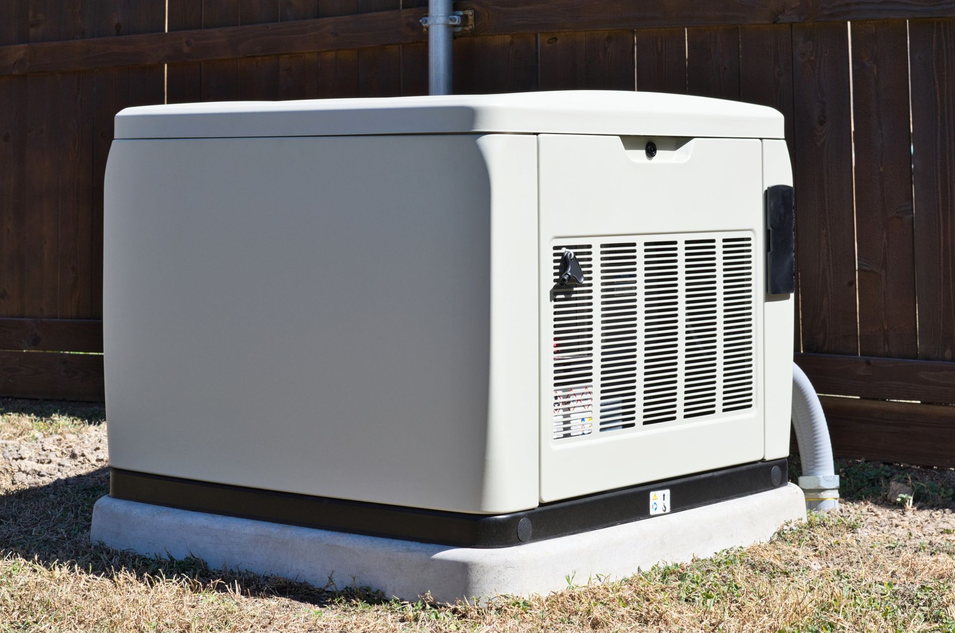 A tan, outdoor standby home generator sitting on a concrete pad in front of a wooden fence.