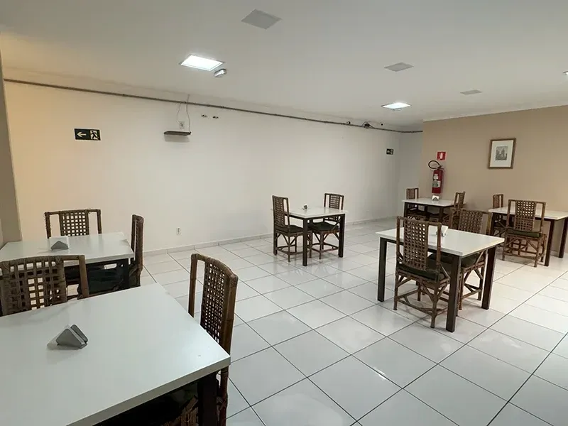 Uma sala de jantar vazia com mesas quadradas brancas e cadeiras de madeira escura sobre um piso de azulejos brancos, iluminada por luzes no teto.