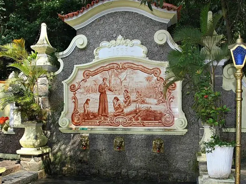 Uma fonte ornamental ao ar livre com um mural de cerâmica em tons de sépia representando uma figura de vestes interagindo com outras pessoas.