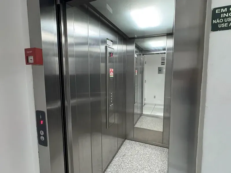 Uma porta de elevador aberta, revelando um interior de aço inoxidável e um espelho na parede do fundo, em um corredor de paredes brancas.