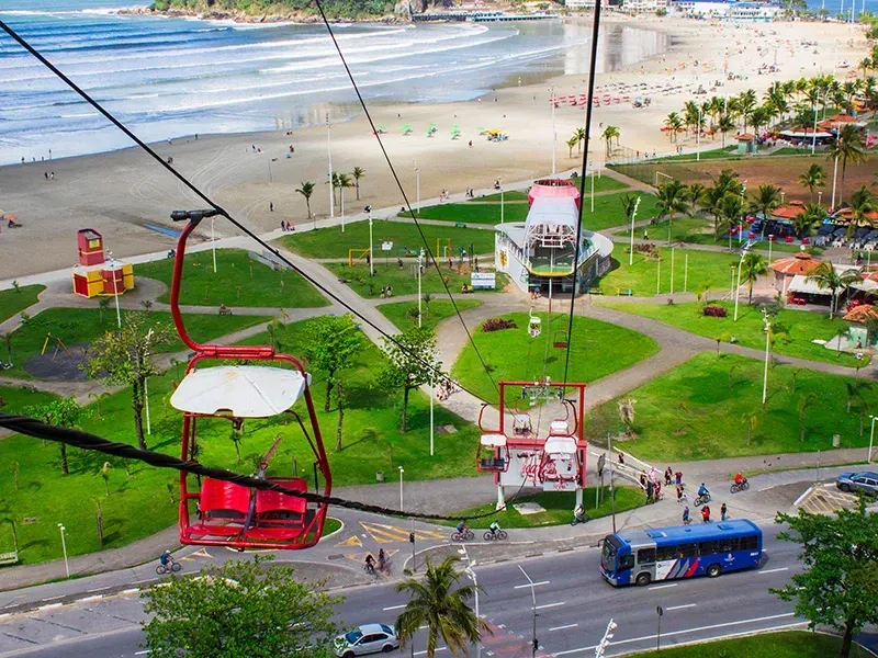Um teleférico vermelho sobre um parque verde, uma rua e uma praia em uma cidade litorânea.