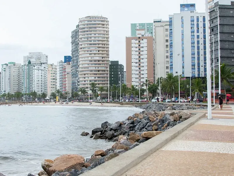 Um calçadão à beira-mar, ladeado por altos prédios de apartamentos e um litoral rochoso, sob um céu nublado.
