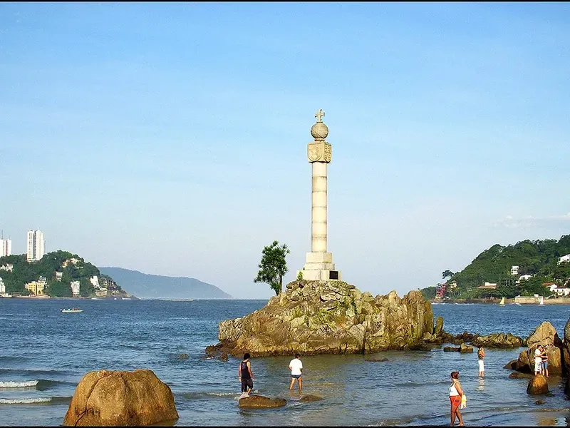 Um monumento de pedra ergue-se no topo de um ilhéu rochoso numa baía, rodeado de água e banhistas sob um céu azul límpido.