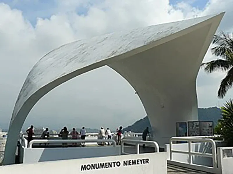 Um monumento branco e curvo de Niemeyer domina o mar, com um grupo de pessoas em um mirante próximo.