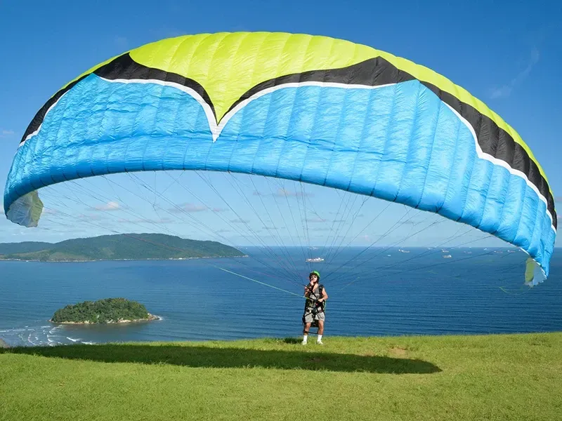 Uma pessoa está em uma colina gramada segurando um grande parapente azul, verde e preto, com vista para o mar e a costa.