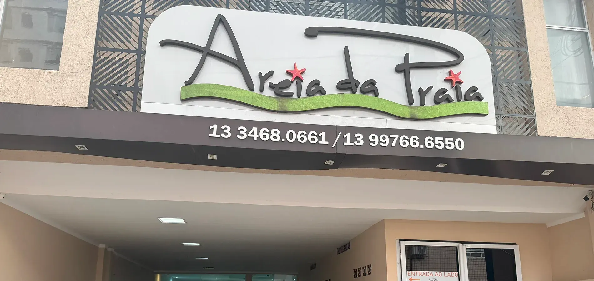 Placa com o nome "Areia da Praia" e números de telefone exibidos na fachada bege do prédio.