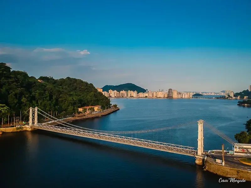 Vista aérea da ponte suspensa Hercilio Luz sobre as águas azuis, com uma encosta verdejante e o horizonte da cidade ao longe.