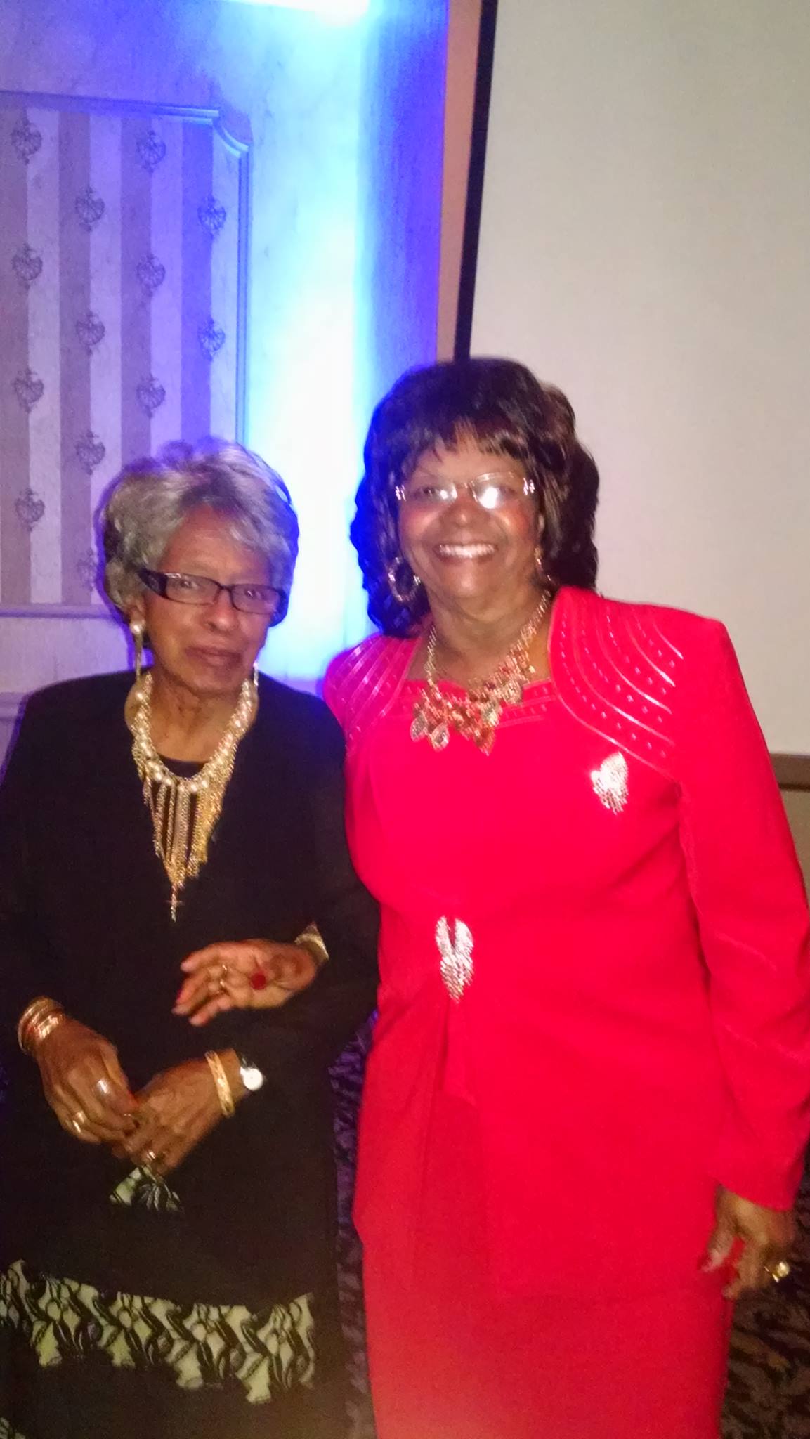 Two women posing together indoors, one in black and gold, the other in a bright red dress