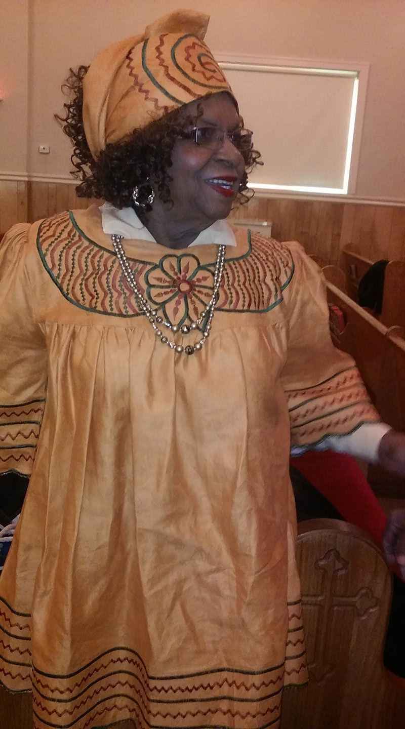 Person in an orange traditional outfit and matching hat, smiling and looking to the side indoors.