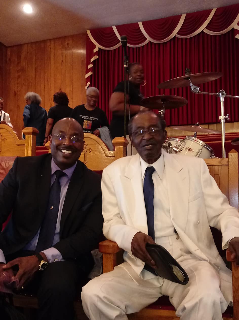 Two men seated in a banquet hall, smiling in suits with red curtains and stage drums behind them