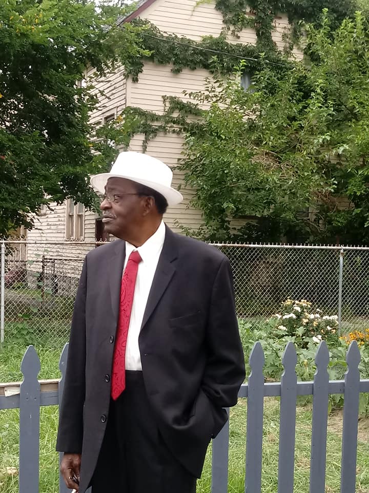 Man in a black suit, red tie, and white hat standing by a picket fence in a yard