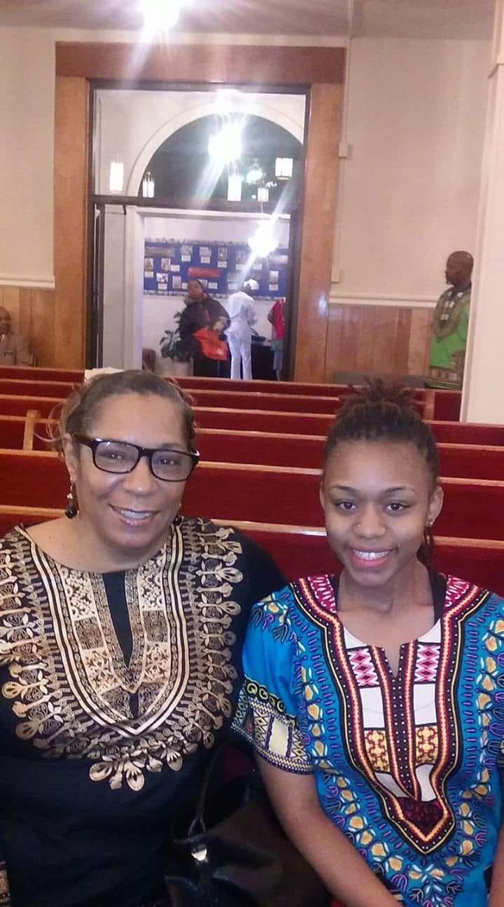 Two people smiling in colorful patterned tops, seated in a church pew with red cushions.