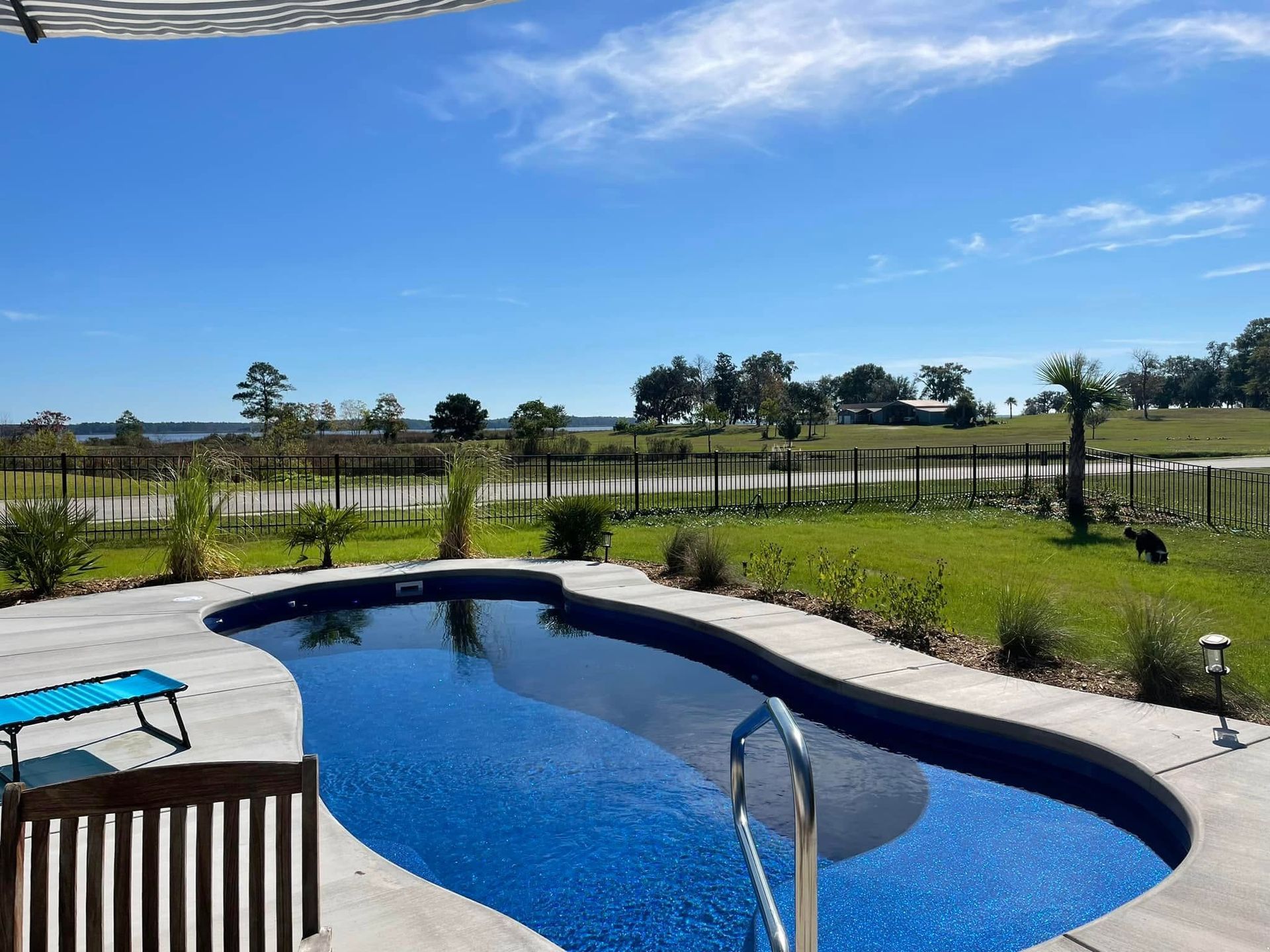 Backyard pool with blue water, patio chairs, lawn, and open field under a clear sky