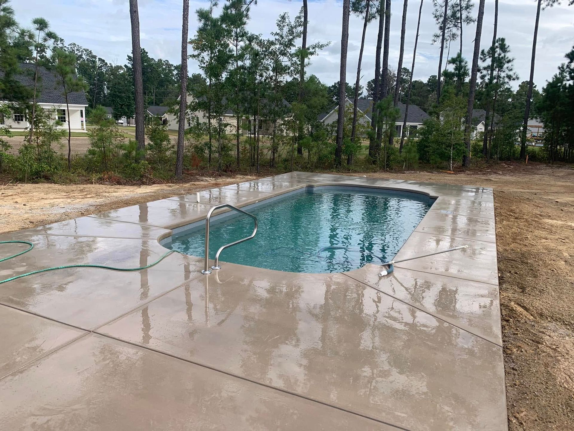Small outdoor swimming pool beside a wet concrete patio, surrounded by trees under an overcast sky