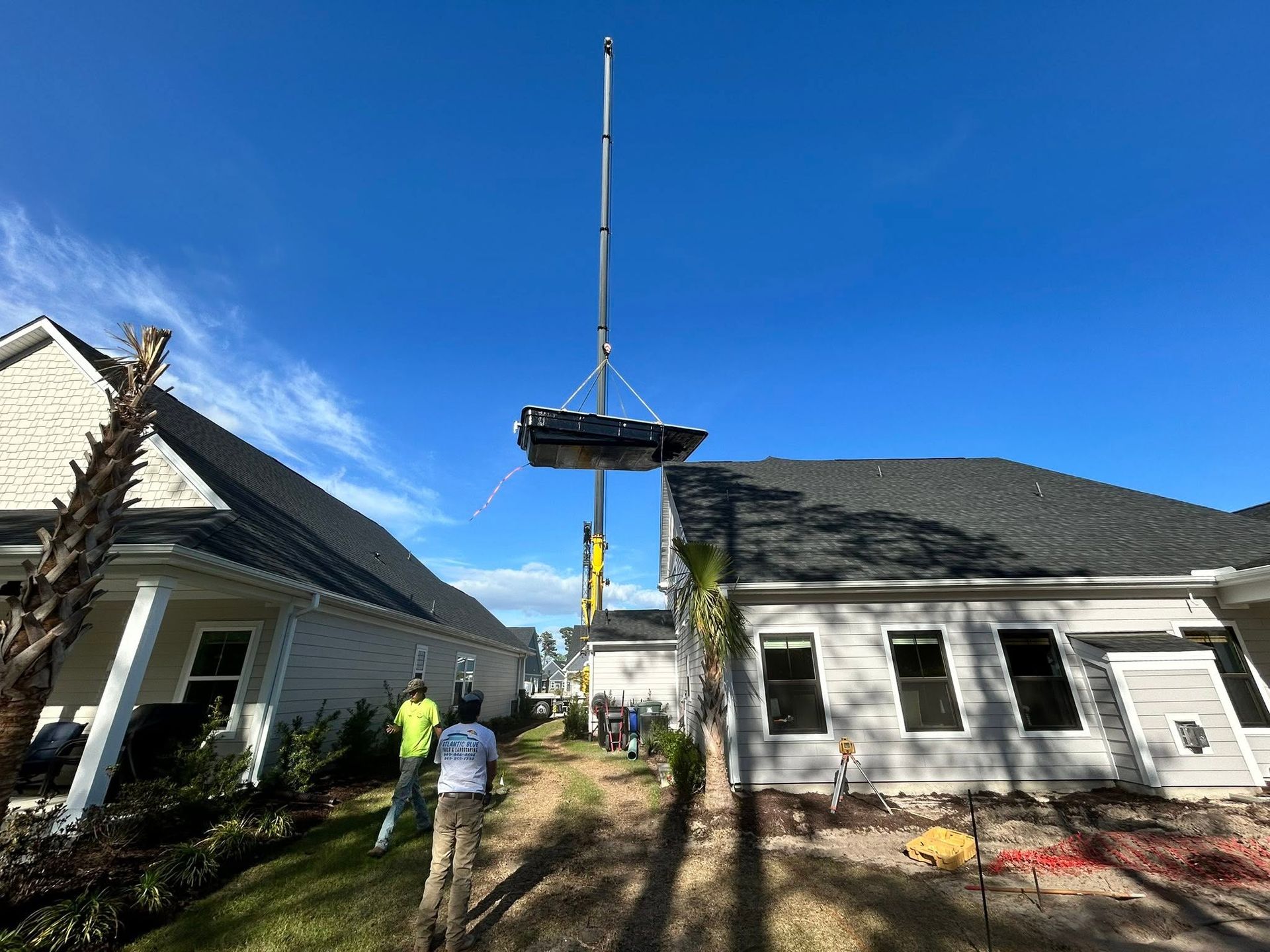 Crane lifting a rooftop section between two houses, with workers below on a sunny day.