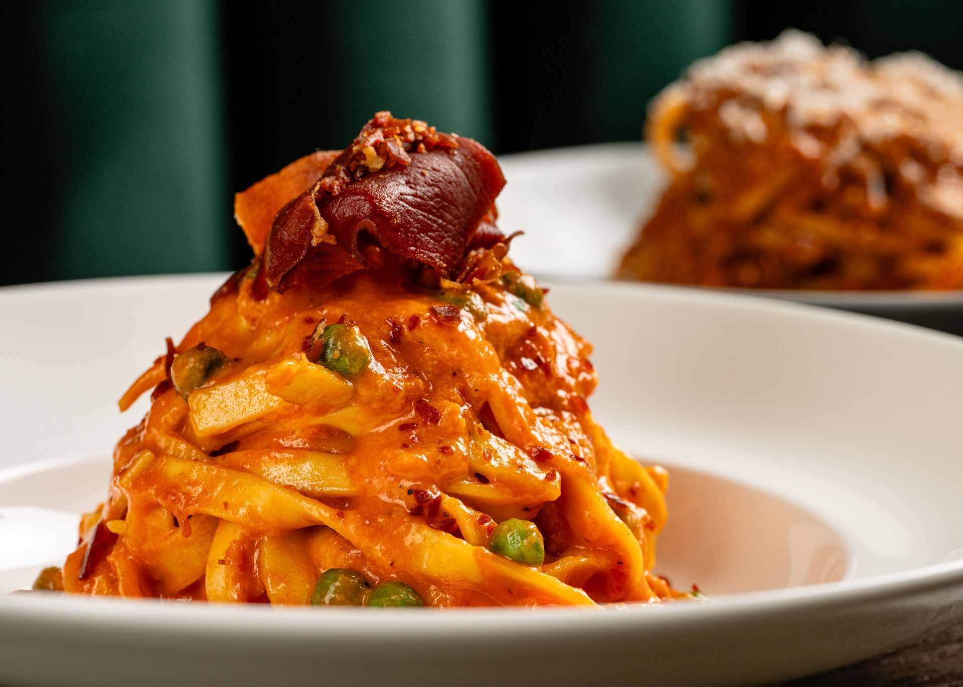 Pasta topped with red sauce and bacon on a white plate, with another blurred dish in the background