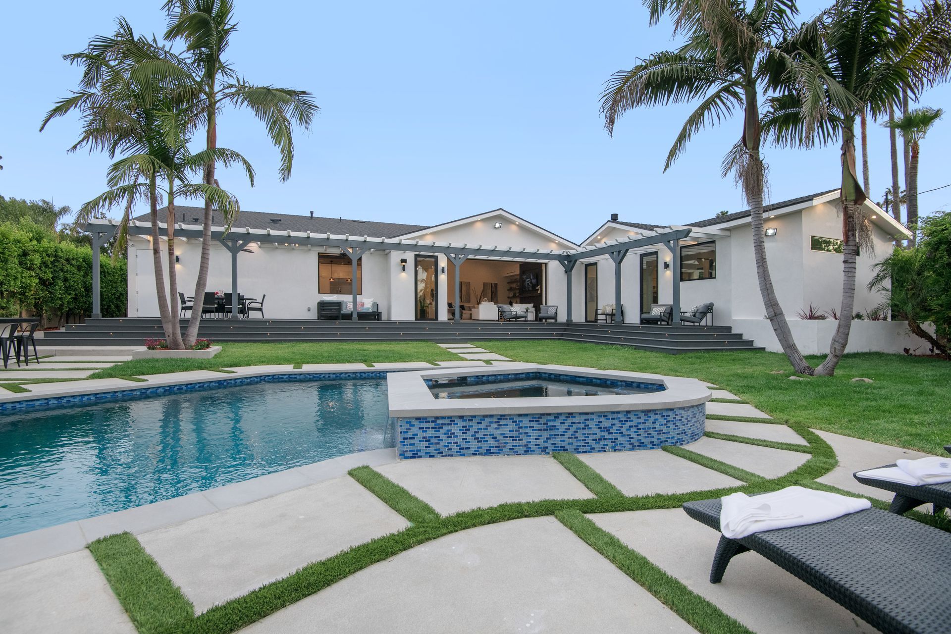 Luxury backyard with a pool, hot tub, palm trees, and a white modern house under a clear sky Luxury backyard with a pool, hot tub, palm trees, and a white modern house under a clear sky