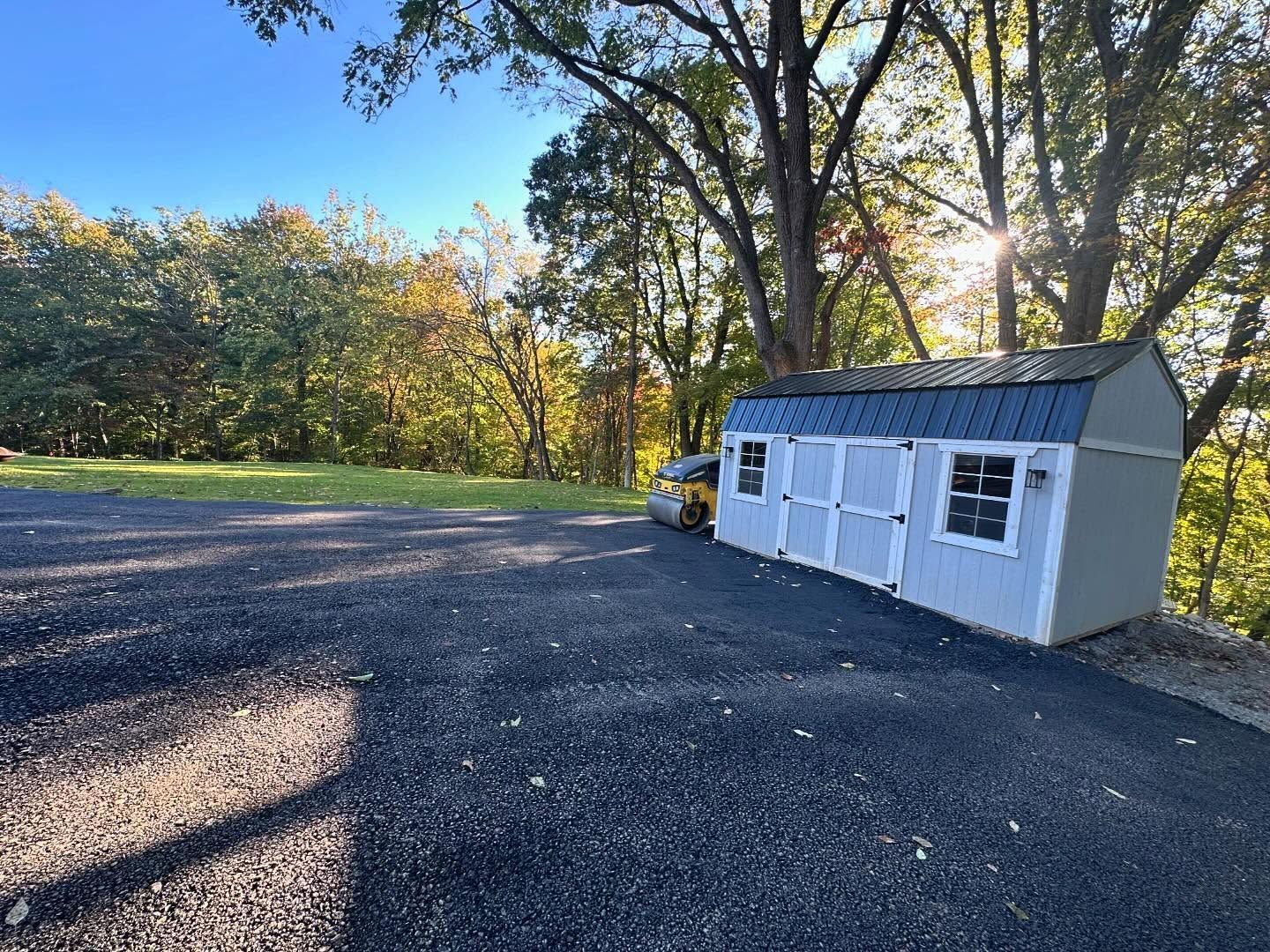 White shed with dark roof beside a gravel driveway, with a yellow car and trees in sunlight.