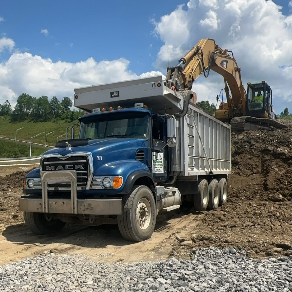 Blue dump truck with raised bed on a dirt construction site beside an excavator on a hillside