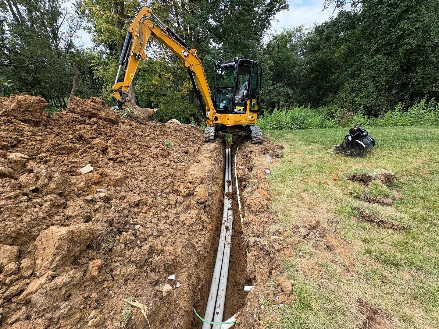 Excavator digging a trench beside a large dirt pile in a grassy field